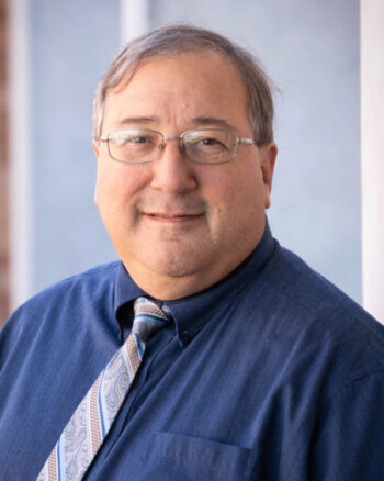 Michael Barber in a blue dress shirt and a patterned tie against a neutral background