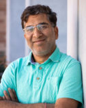 Ramesh Goel in glasses wearing a light blue polo shirt standing against a neutral background